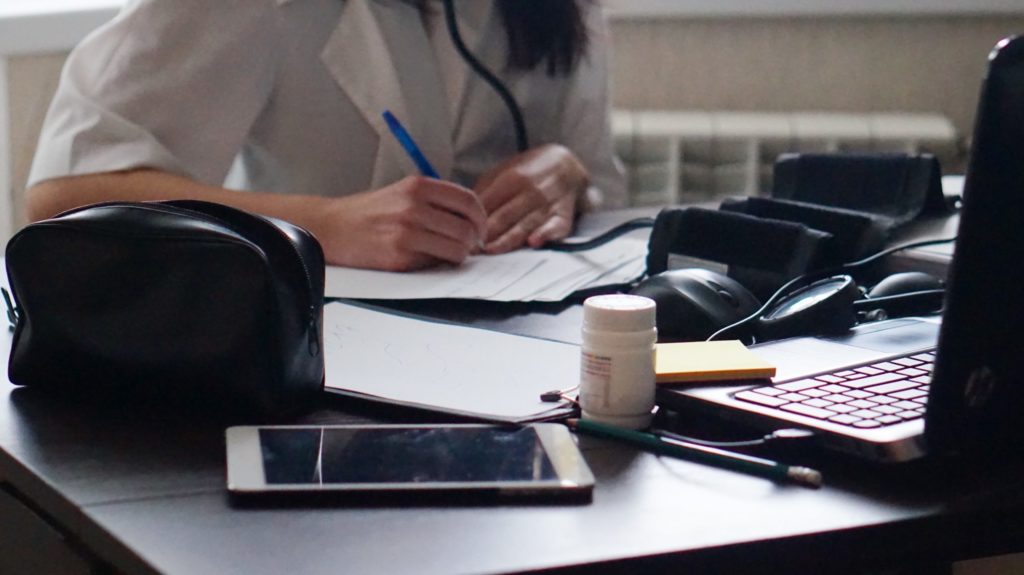 Close-up of a healthcare professional’s hands at a desk with a stethoscope, laptop, and medication bottle, representing a clinical workspace.
